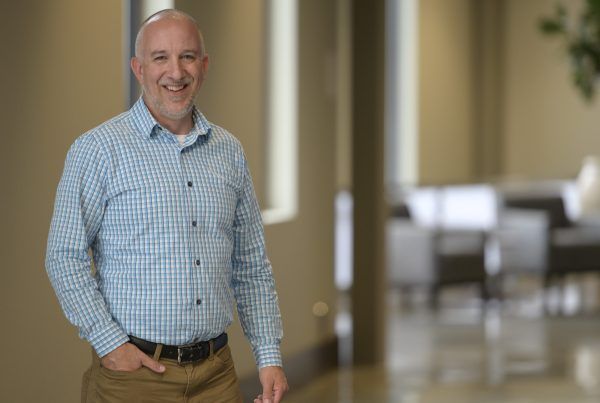 Smiling man in a blue checkered shirt stands in a bright office hallway