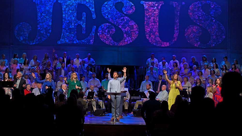 Choir on stage under blue lighting with huge “JESUS” backdrop, singer at center podium