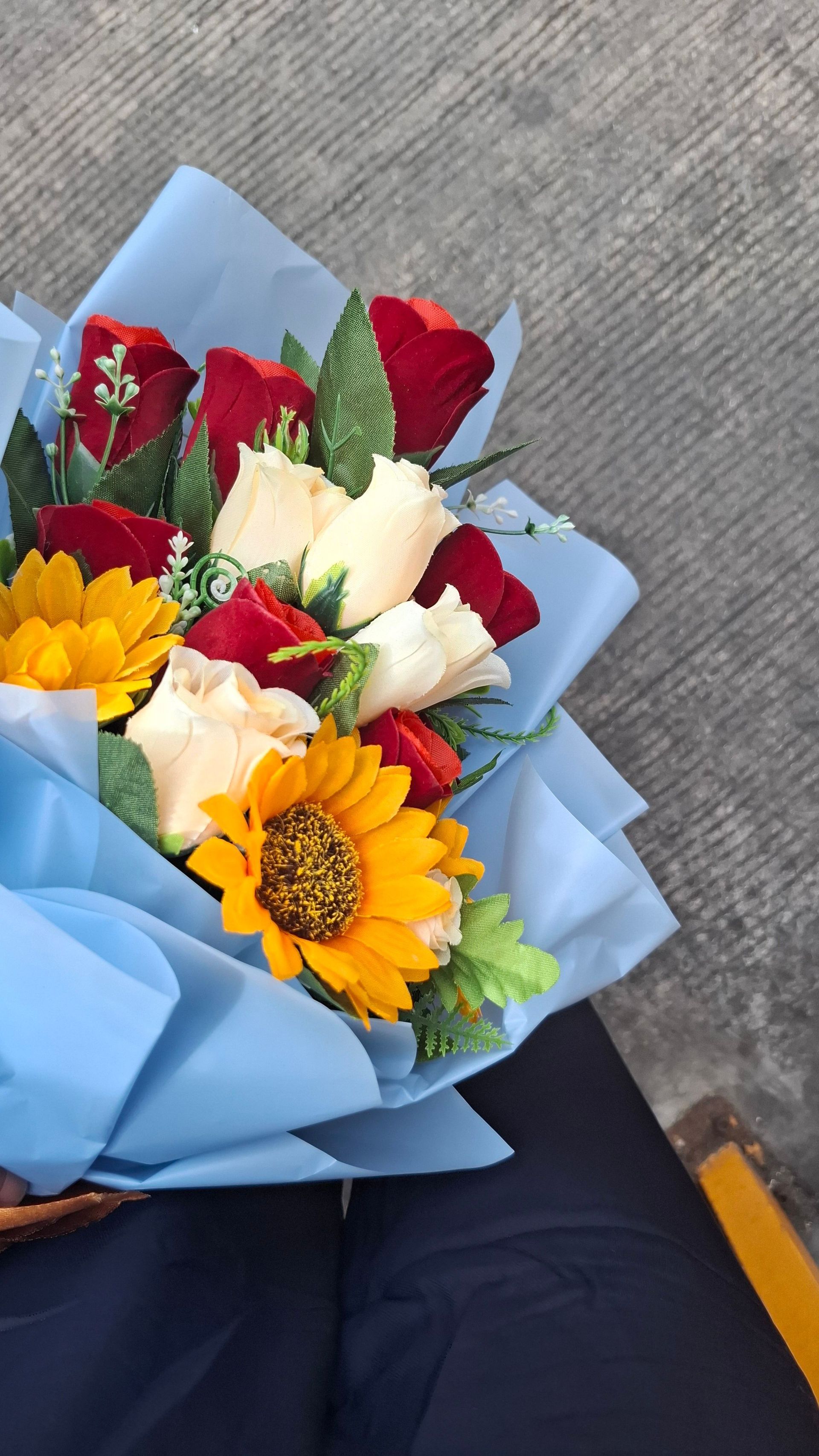 Bouquet of red, white, and yellow flowers wrapped in light blue paper, held outdoors on gray pavement
