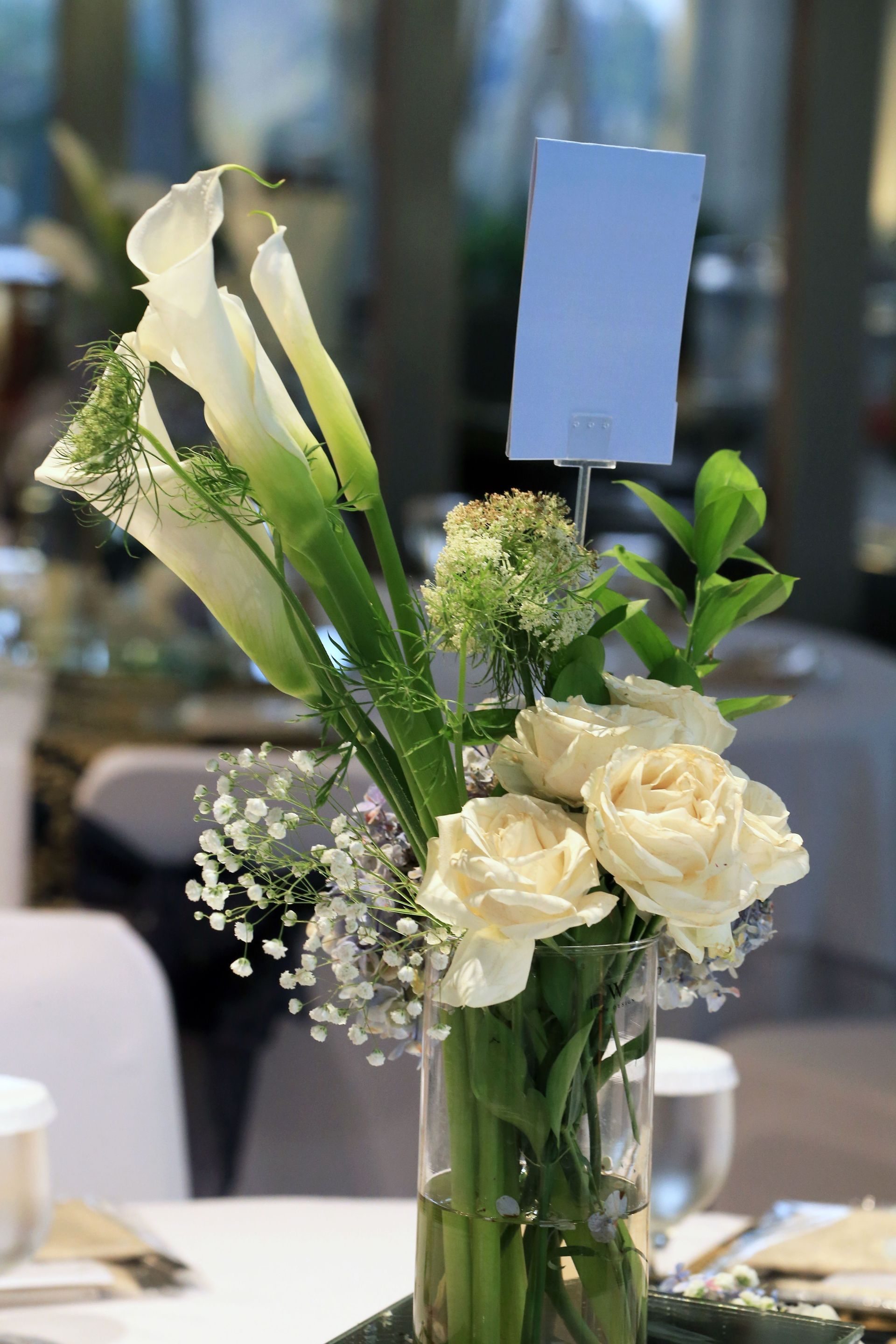 White floral centerpiece with calla lilies and roses in a glass vase on a table, with a blank card on top