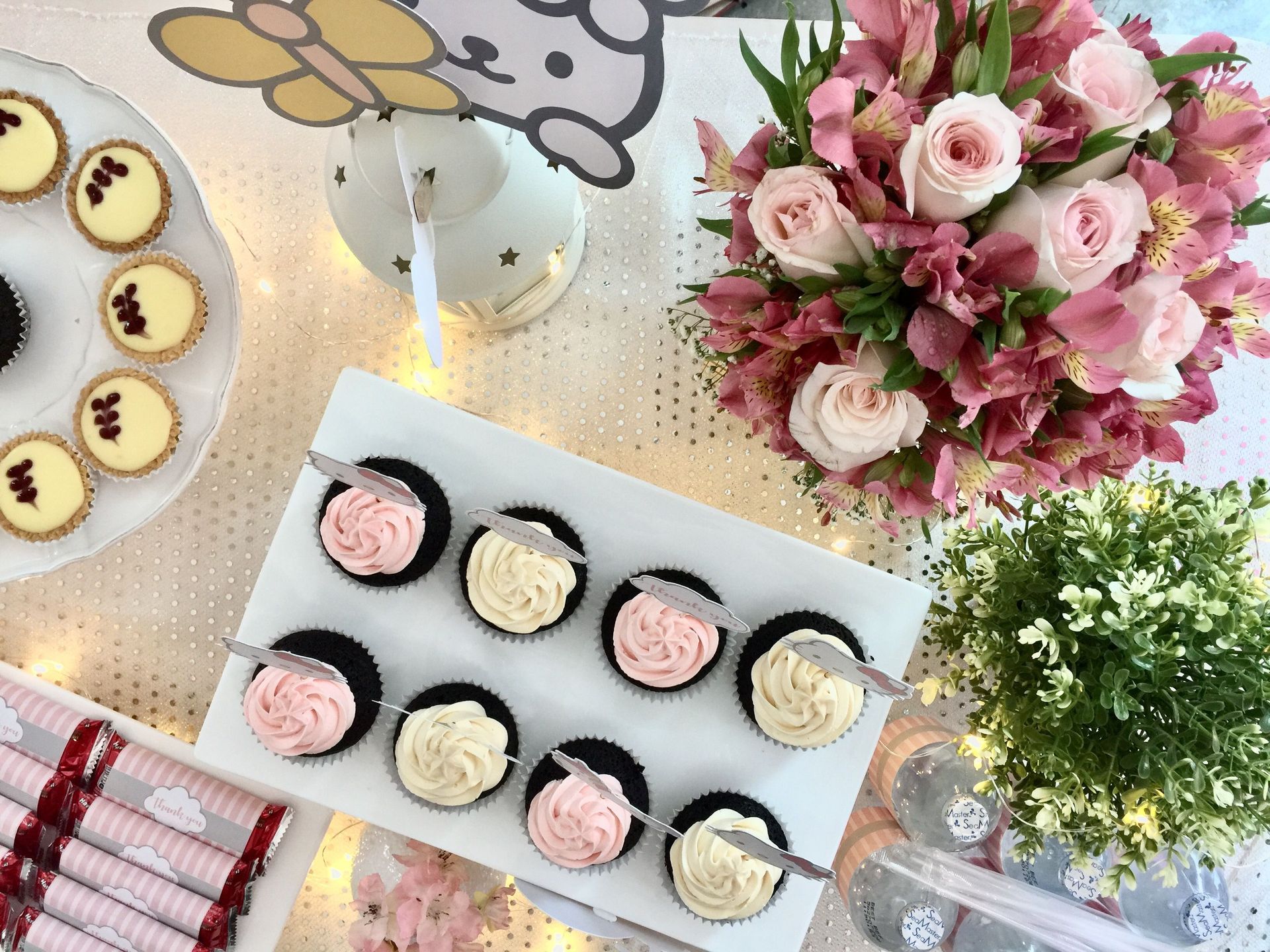 Desserts and flowers on a table, with cupcake trays, a pink rose bouquet, and teacups in a pastel setting