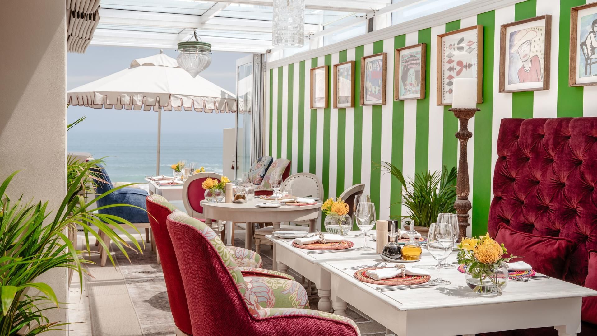 Elegant outdoor café with red velvet seating, white tables, and green-striped wall by the seaside