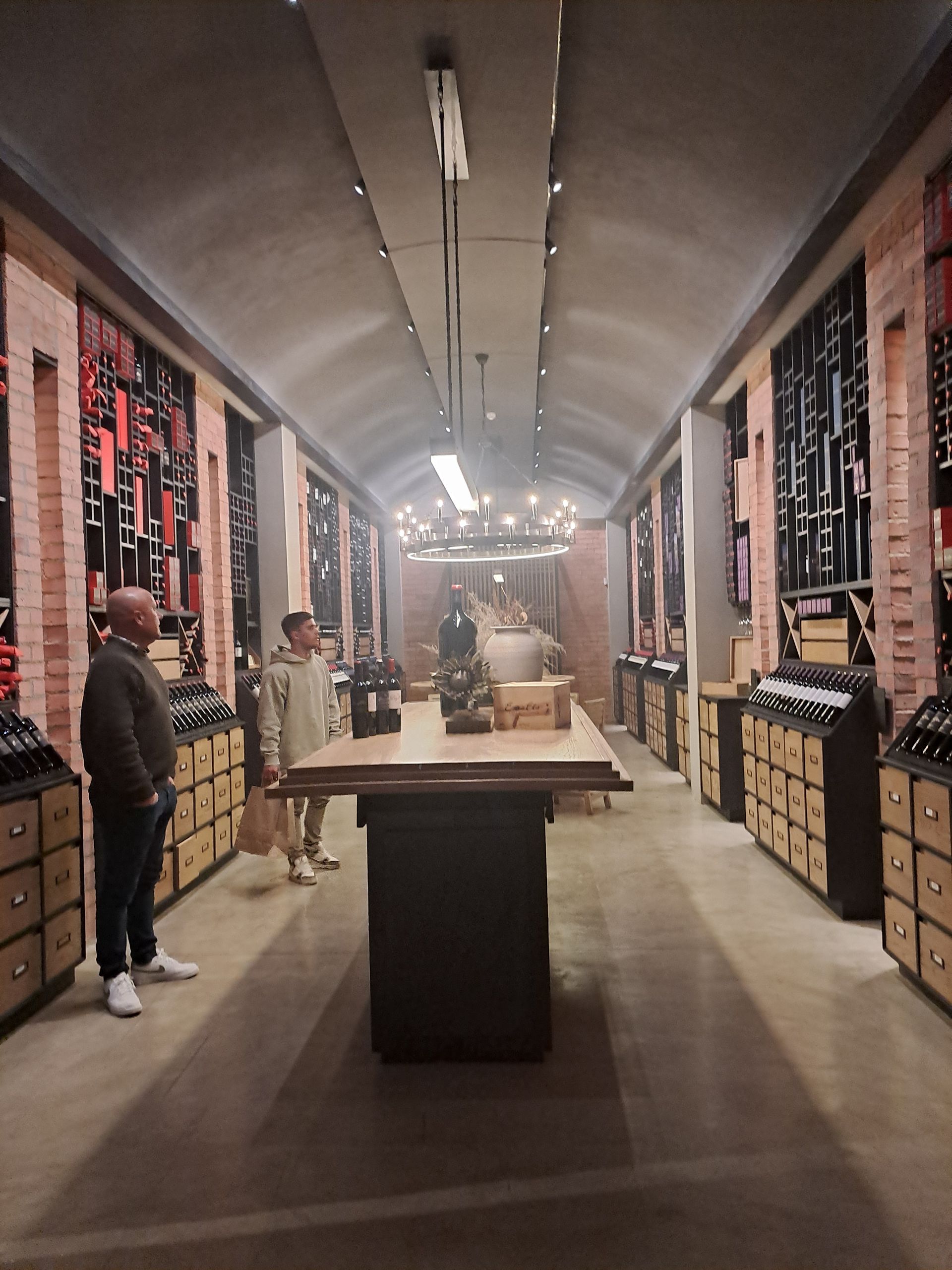Wine shop aisle with racks of bottles on both sides and a few customers browsing.
