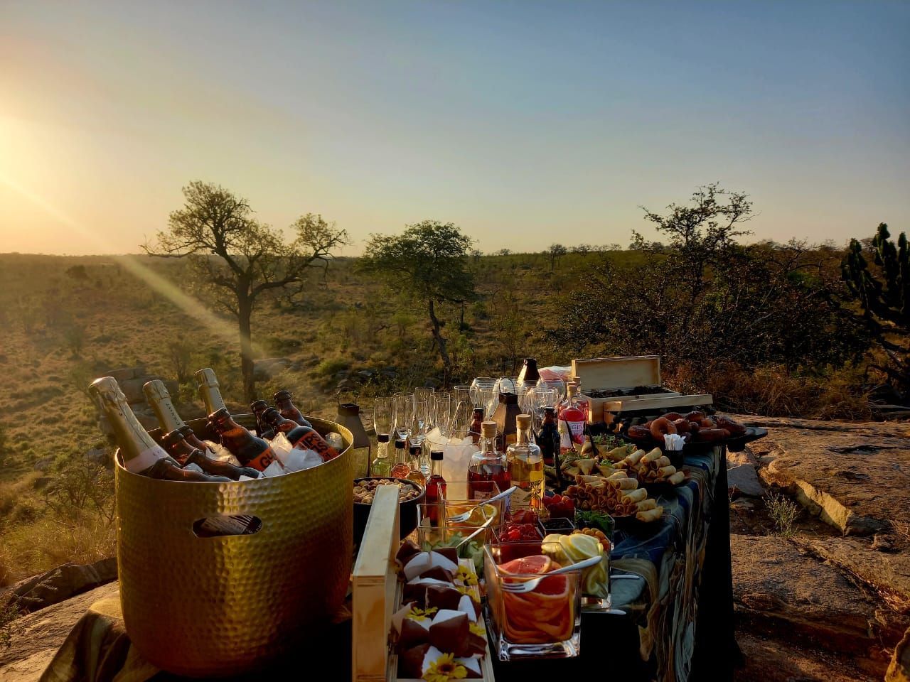 Outdoor picnic spread with drinks and food on a table beside a large ice bucket at sunset in a dry field