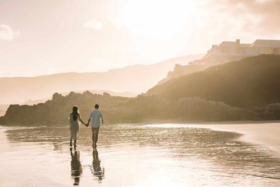 Two people holding hands on a beach at sunset, reflected in wet sand with hills in the background