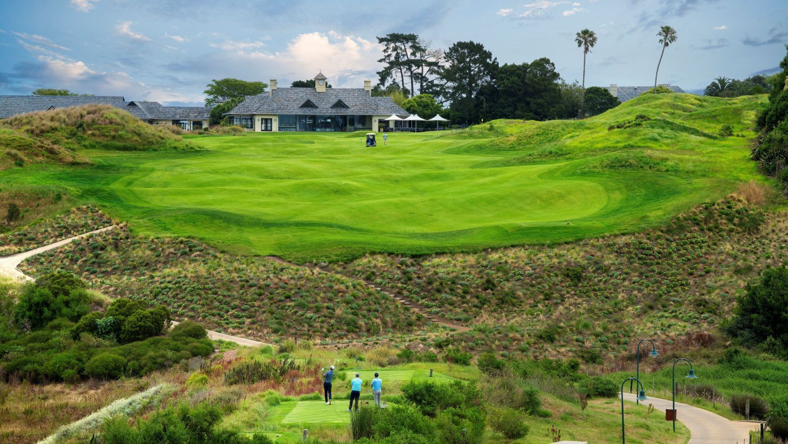A lush golf course fairway with a clubhouse in the distance and two people at the bottom of a hill.