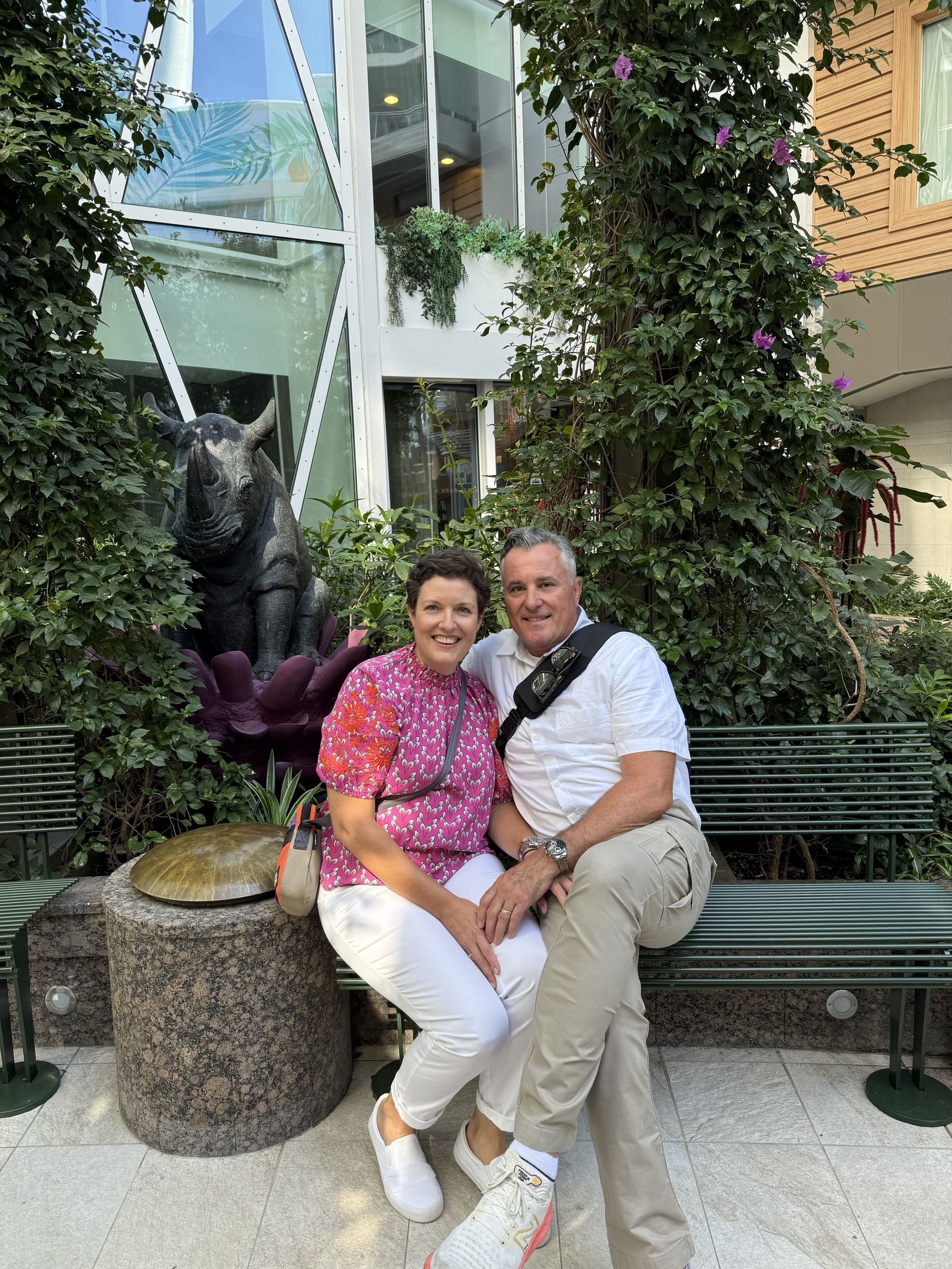 Two people sit on a bench by a fountain and sculpture in a garden setting, smiling at the camera.