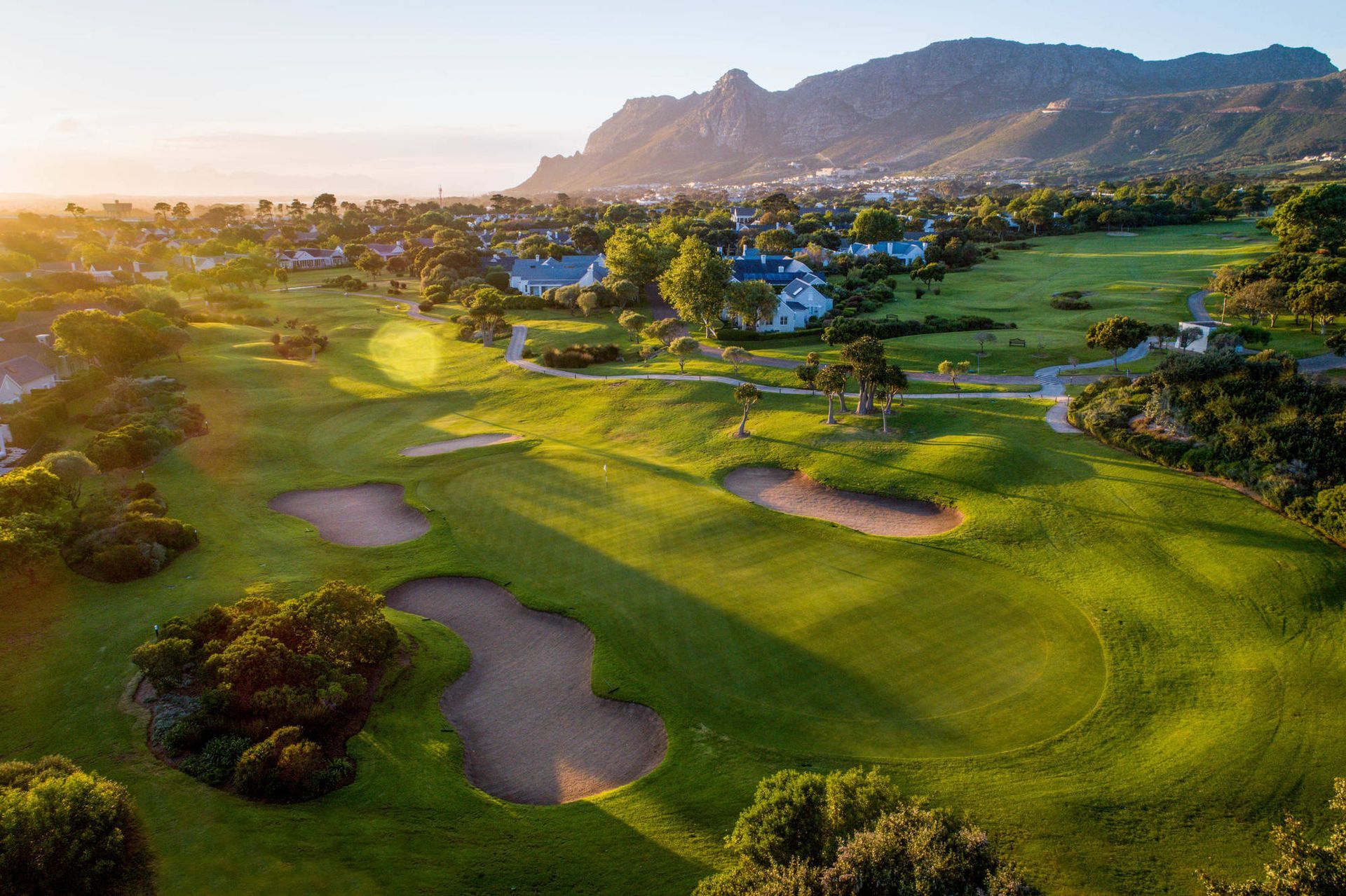Aerial view of a lush green golf course with sand bunkers, palm trees, and mountains at sunset