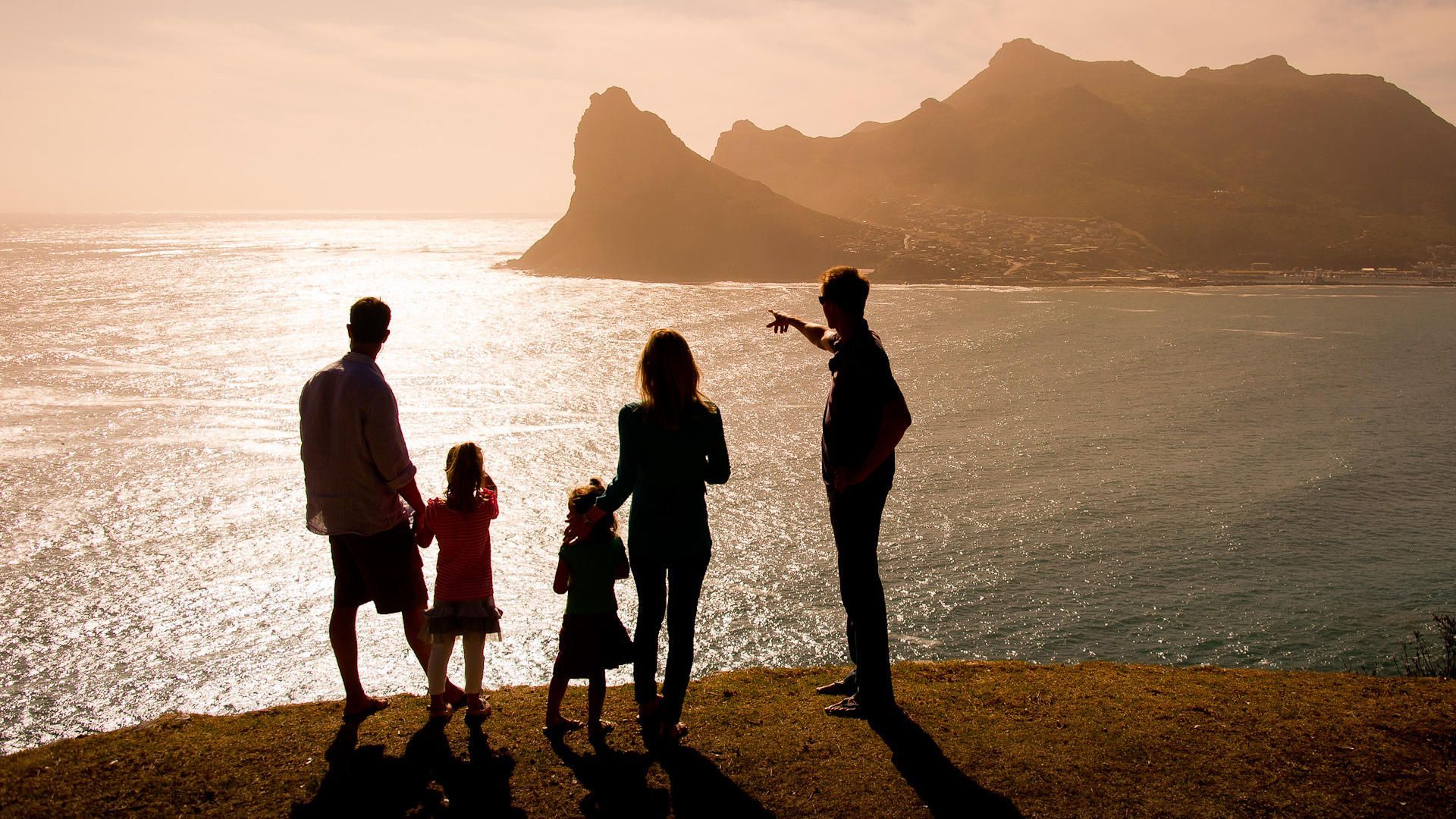 Silhouettes of four people on a grassy cliff, watching the sun set over the ocean and distant mountains.