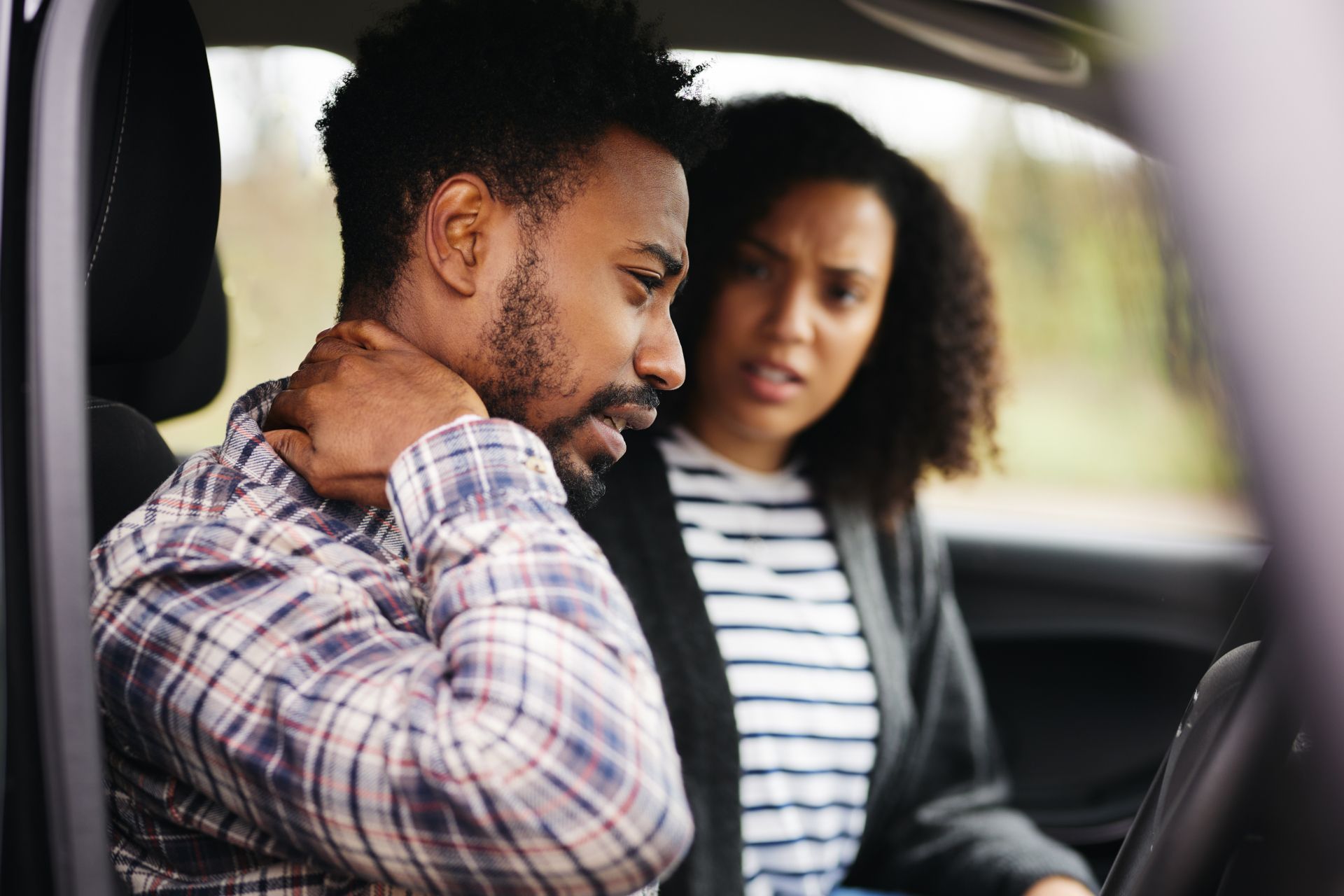 Two people sit in a car, one holding their neck while the other looks at them with concern.