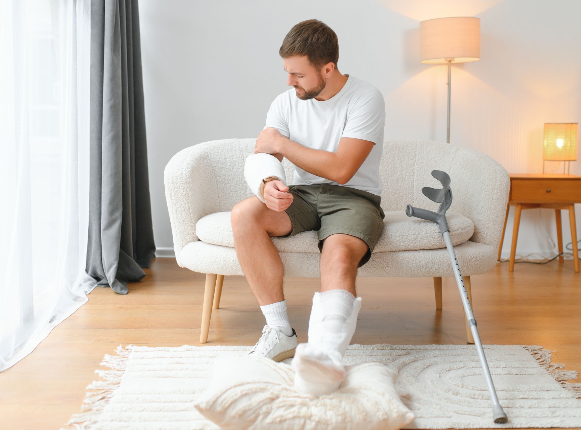 Man in a white T-shirt and shorts rubbing his knee while seated on a sofa in a bright living room, using crutches nearby