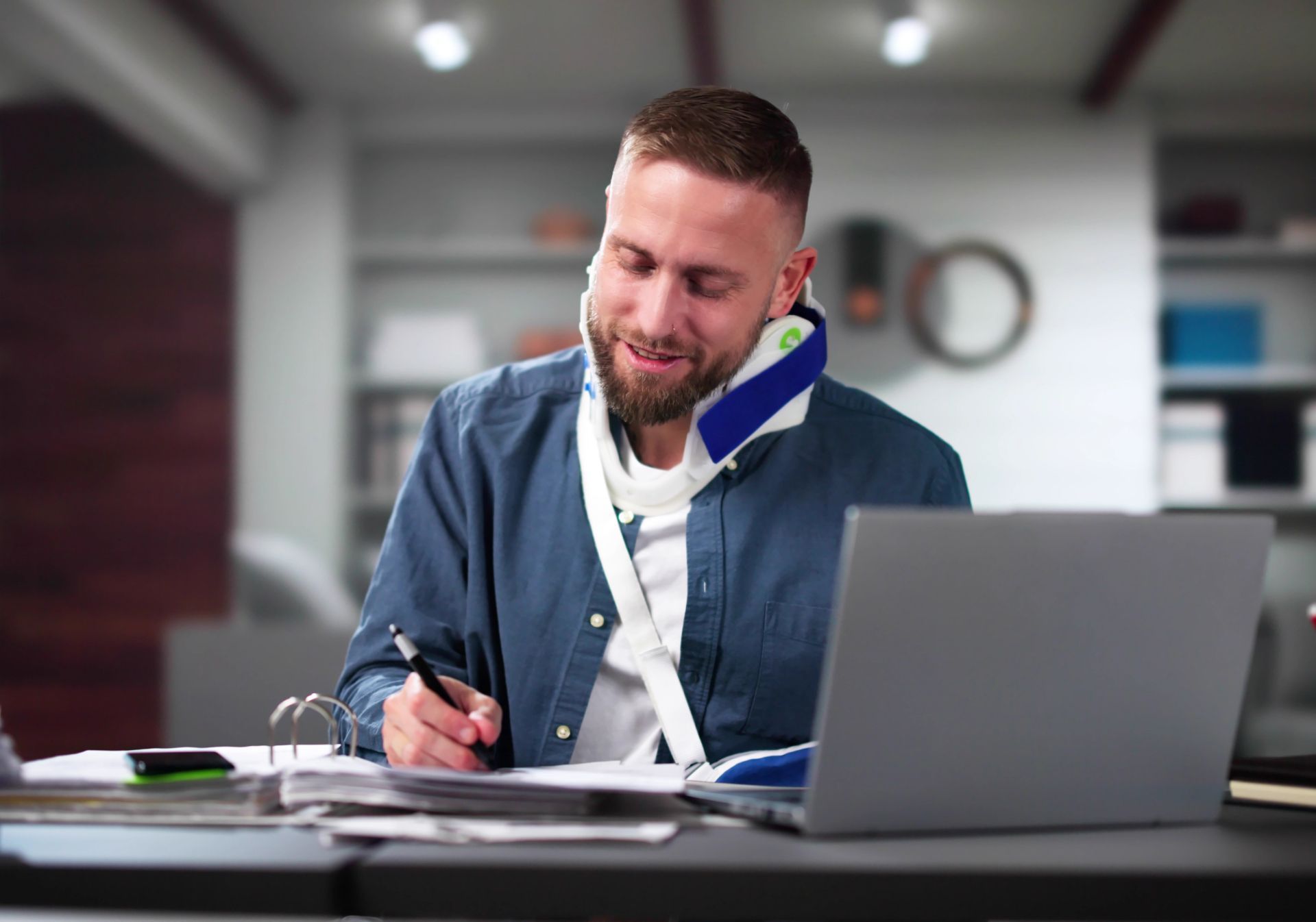 Man with arm in a sling writing in a notebook beside a laptop at a desk