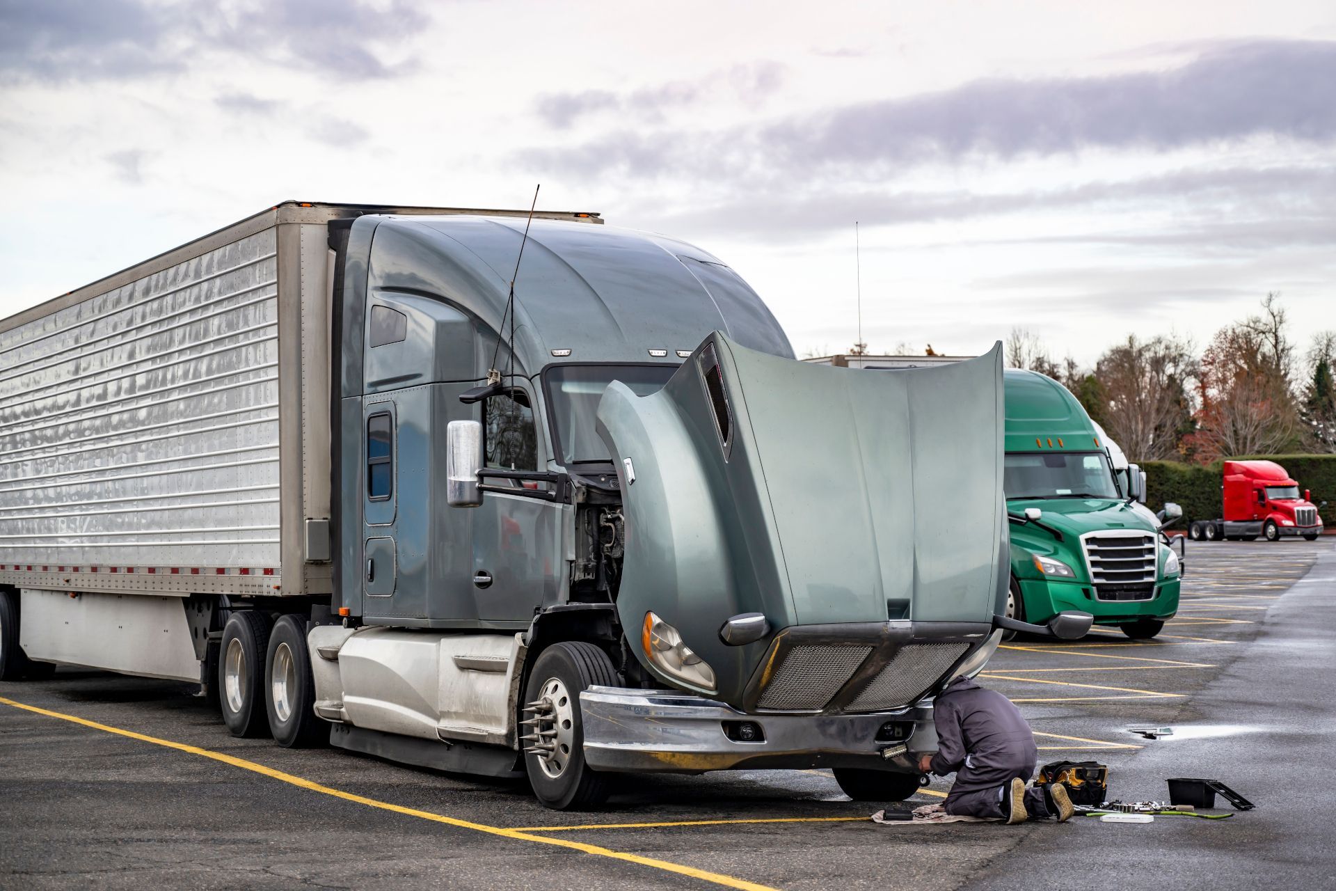 Semi-truck with damaged front hood, a person kneeling beside it in a parking lot.