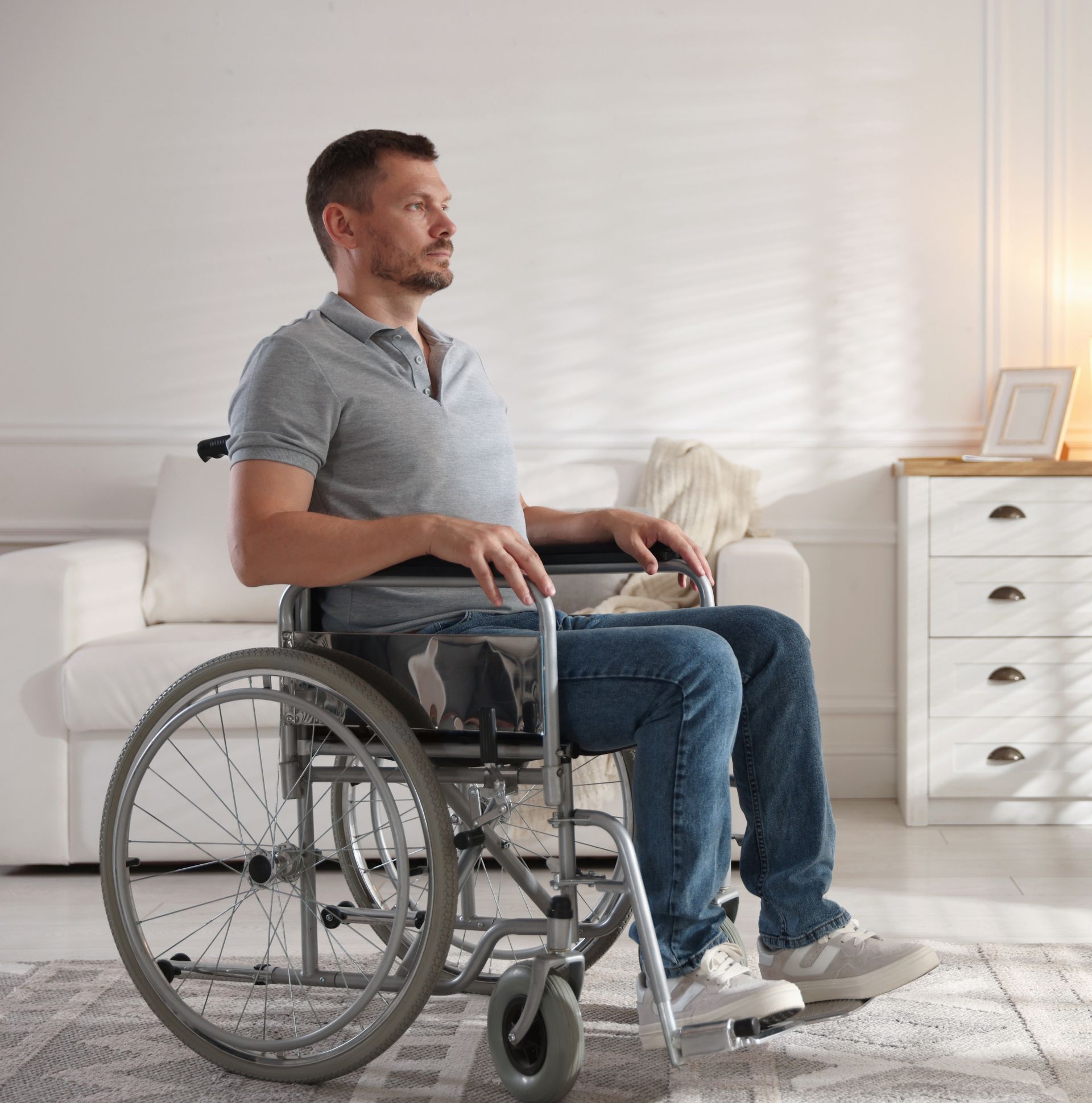 Man seated in a wheelchair in a bright living room, wearing a gray shirt and jeans.