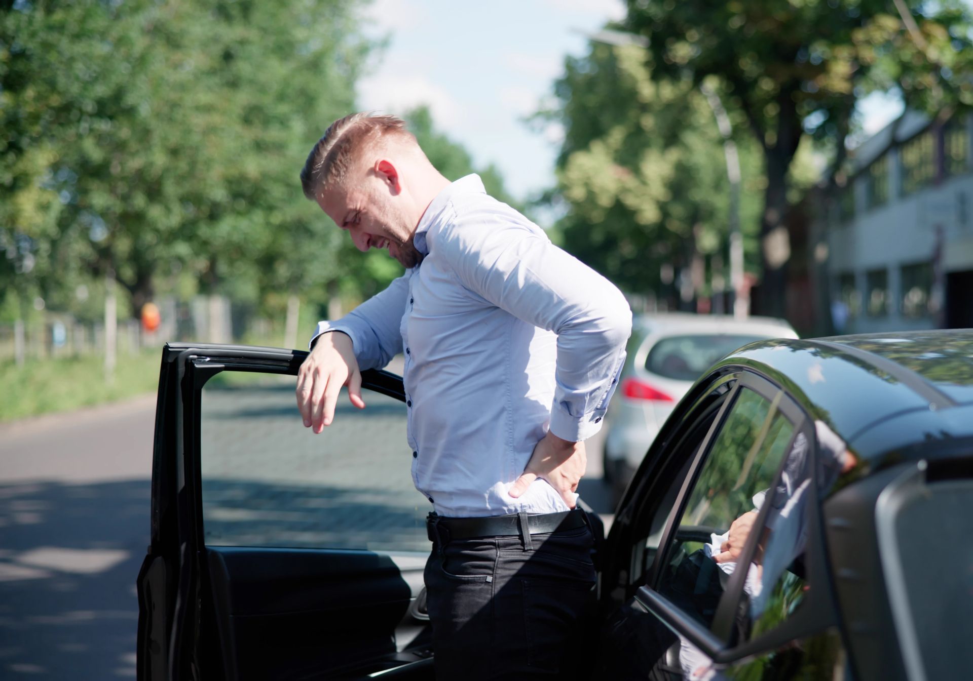 Man standing beside an open car door on a sunny street, leaning into the vehicle
