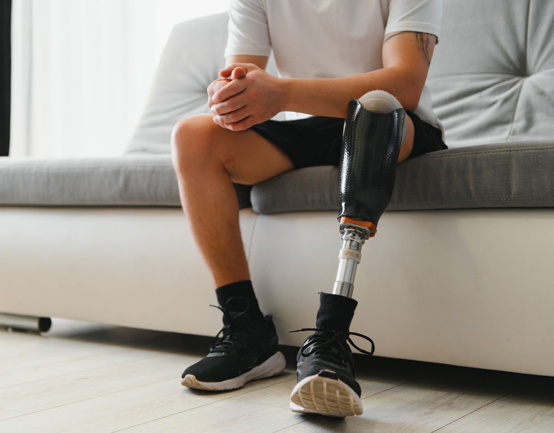 Person seated on a gray couch with a black prosthetic leg and sneakers in a bright living room