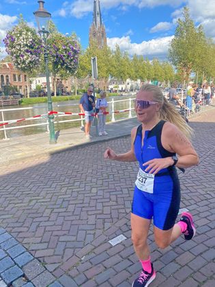 Een vrouw rent in blauwe sportkleding en roze schoenen over een pad langs een kanaal onder een zonnige hemel.