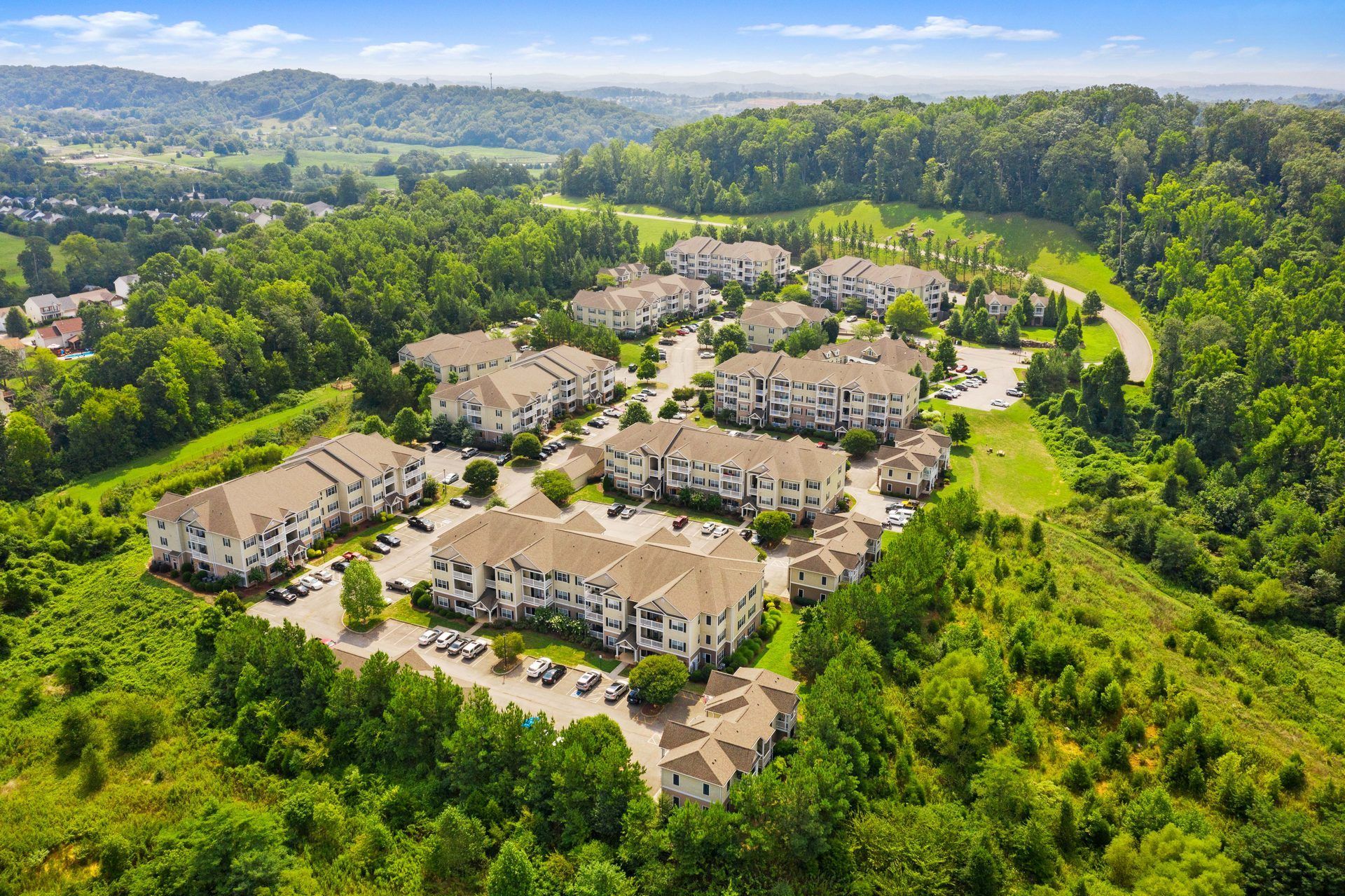 Aerial view of apartment buildings nestled in a lush green valley with parking lots and surrounding trees under a bright blue sky.