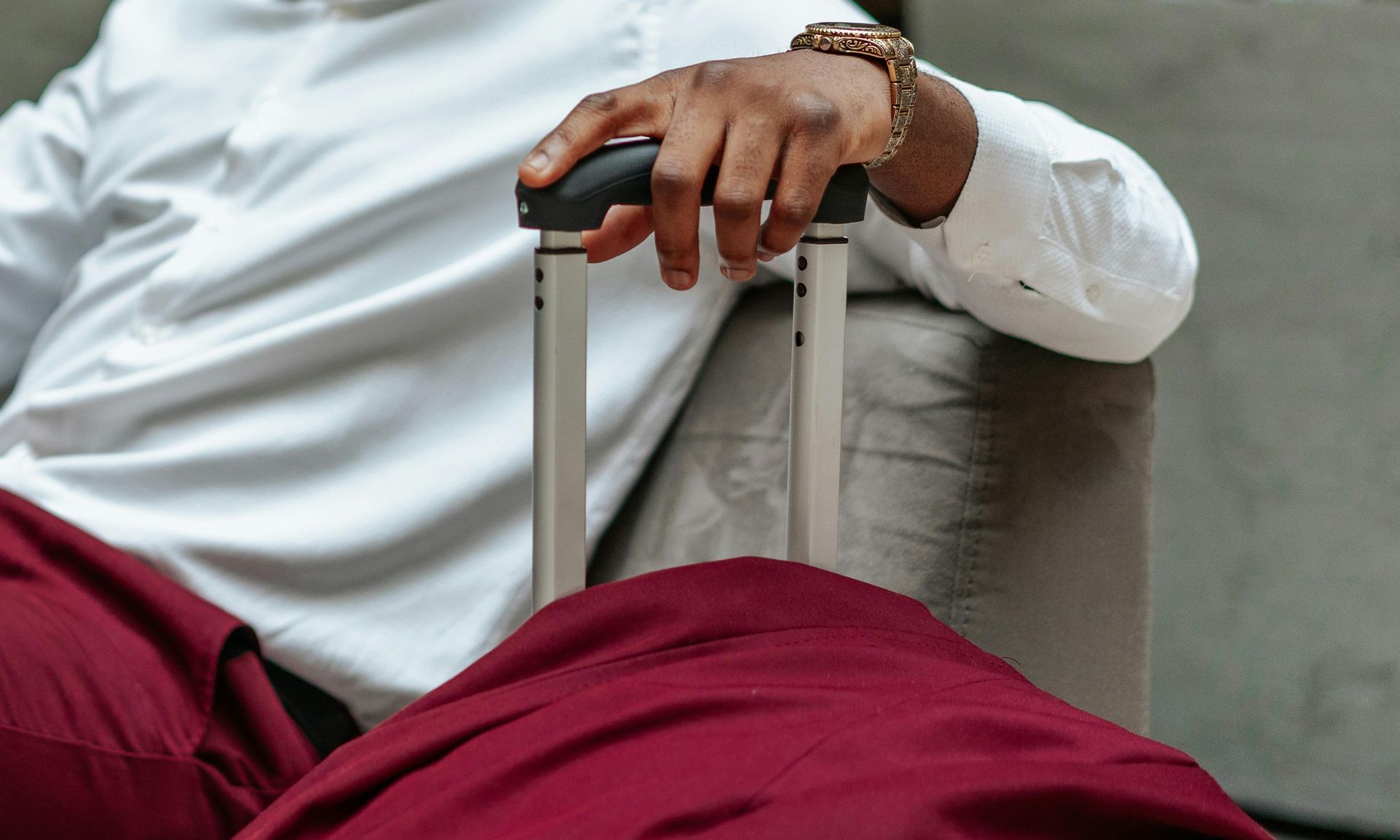Person with luggage, sitting. Hand holding handle, wearing white shirt, red pants.