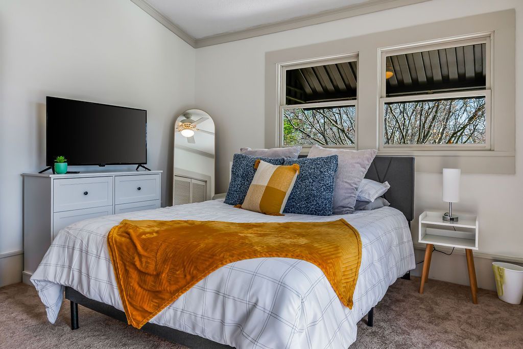 Bedroom with bed, dresser, TV, mirror, and two windows. Yellow blanket and blue pillows. White walls.