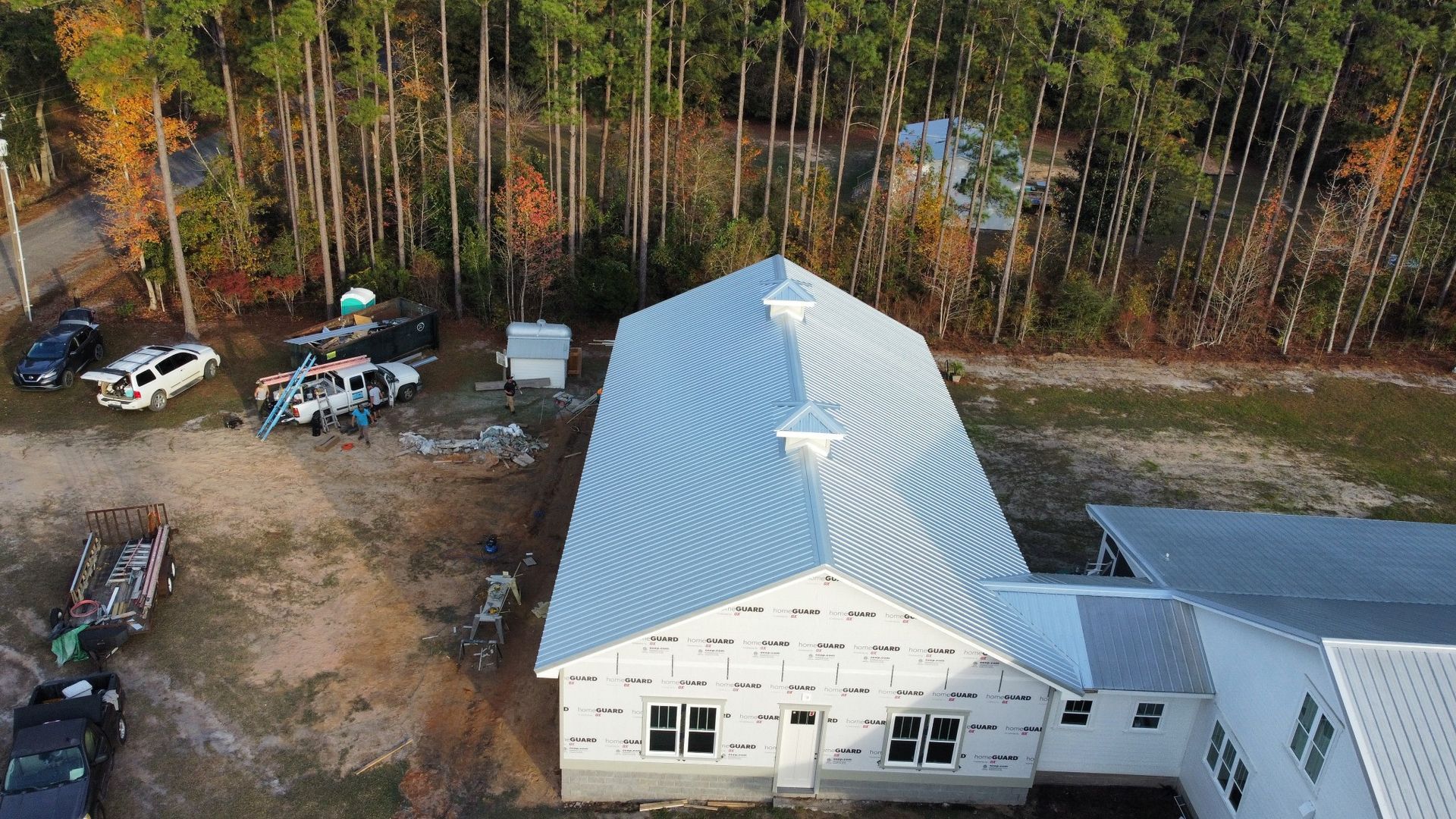 Aerial view of a white building with a metal roof beside a wooded area and parked vehicles