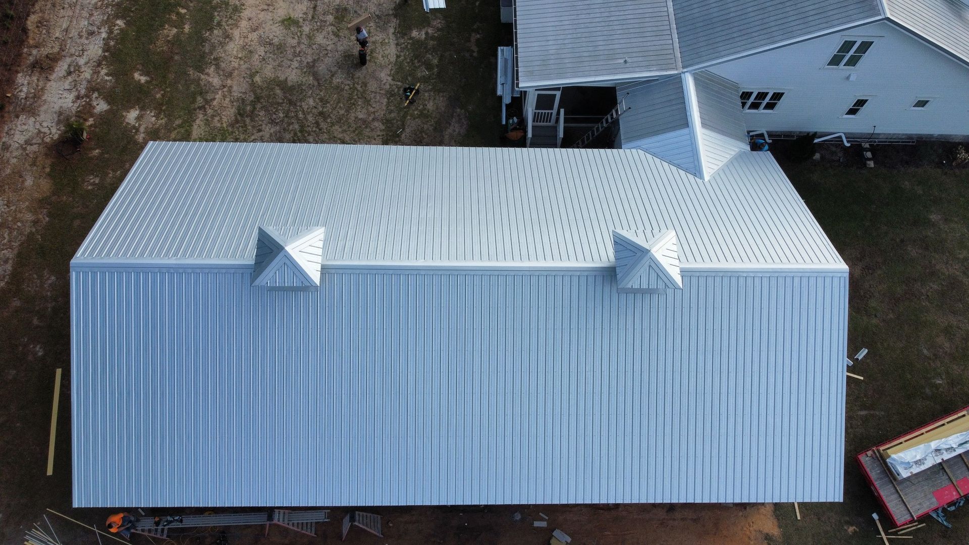 Aerial view of a silver metal roof with two small vents beside a white house and trees.