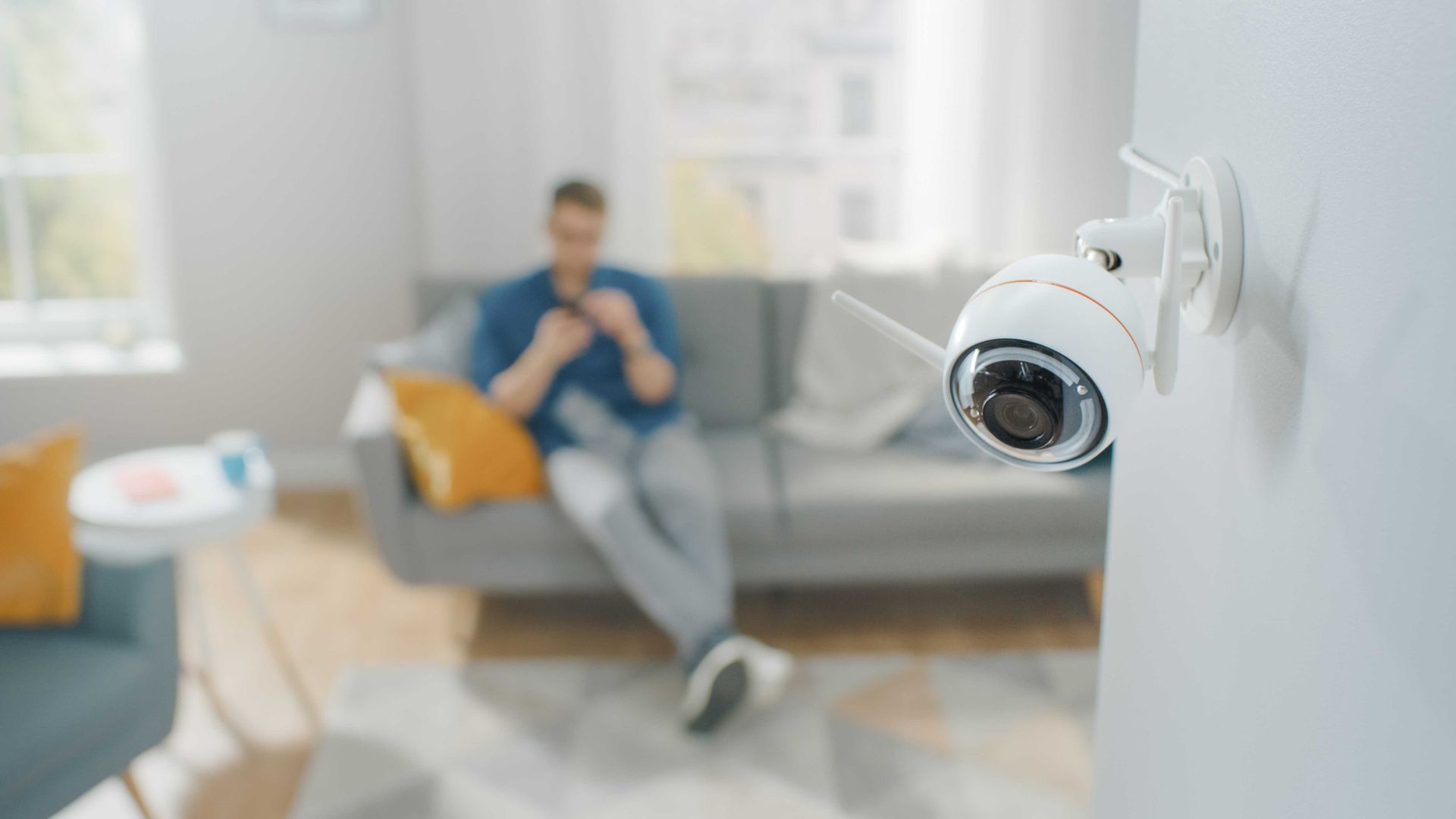 Caméra de sécurité fixée au mur, observant un salon où une personne utilise son téléphone sur un canapé.