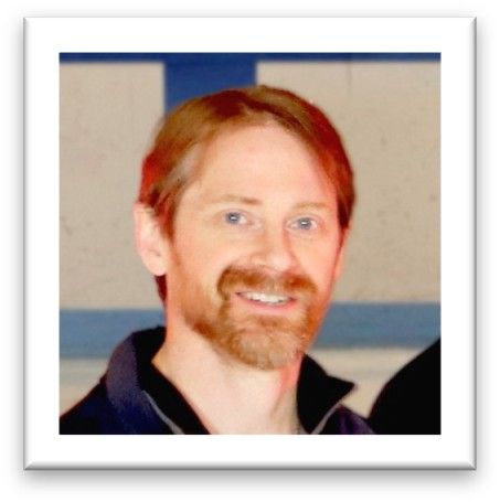 Portrait of a smiling person with red hair and a beard, wearing a dark collared shirt, against a blurred indoor background