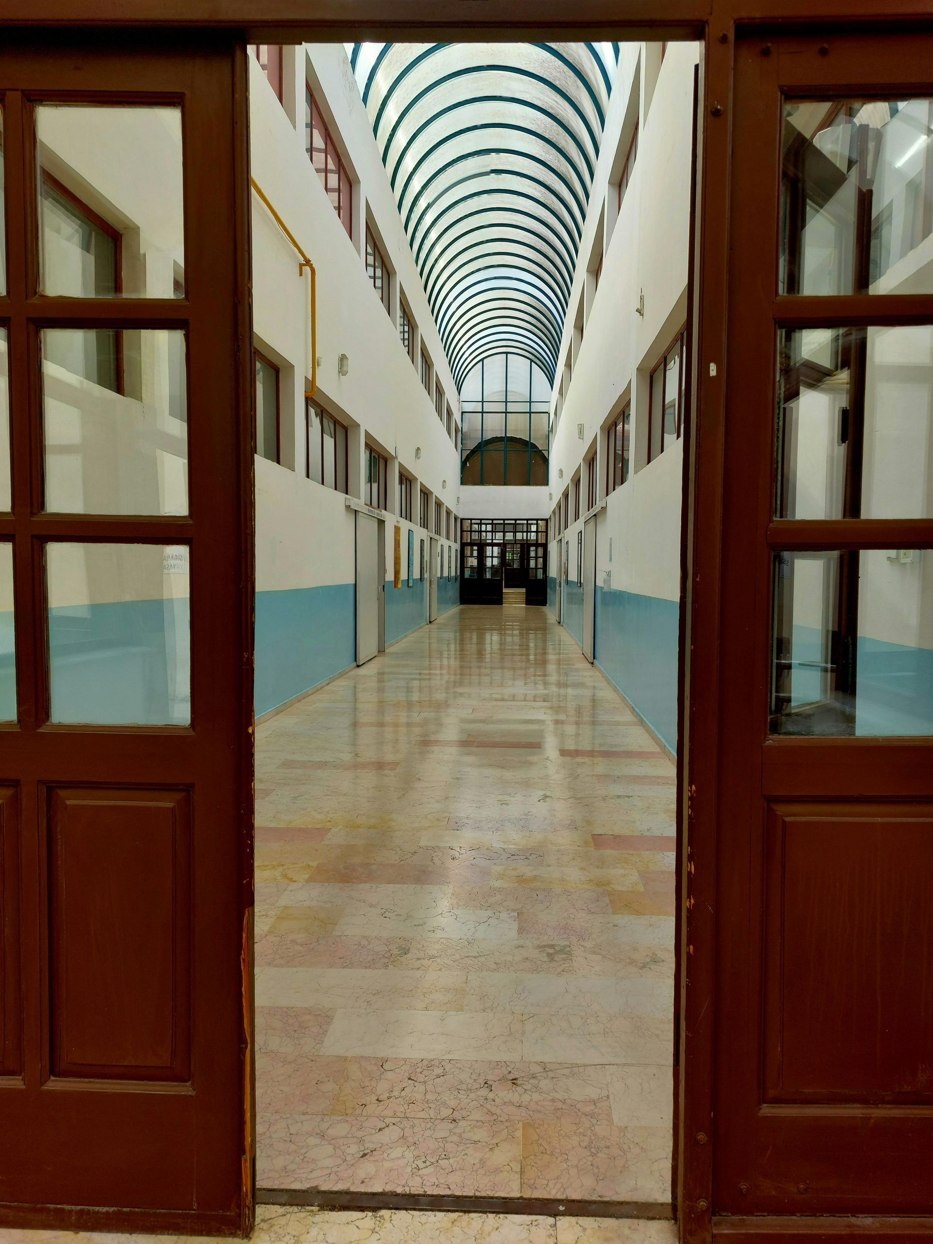 Long hallway with arched glass ceiling and open wooden doors, leading to a distant doorway.