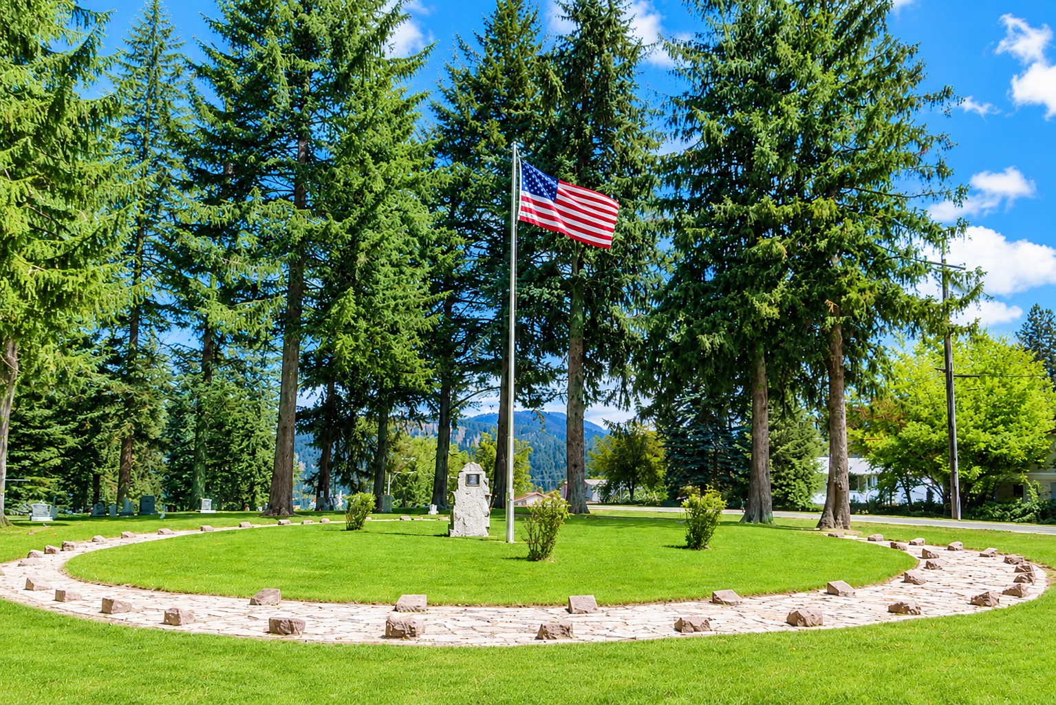 American flag on a pole in a landscaped park with tall evergreen trees and a grassy circular bed
