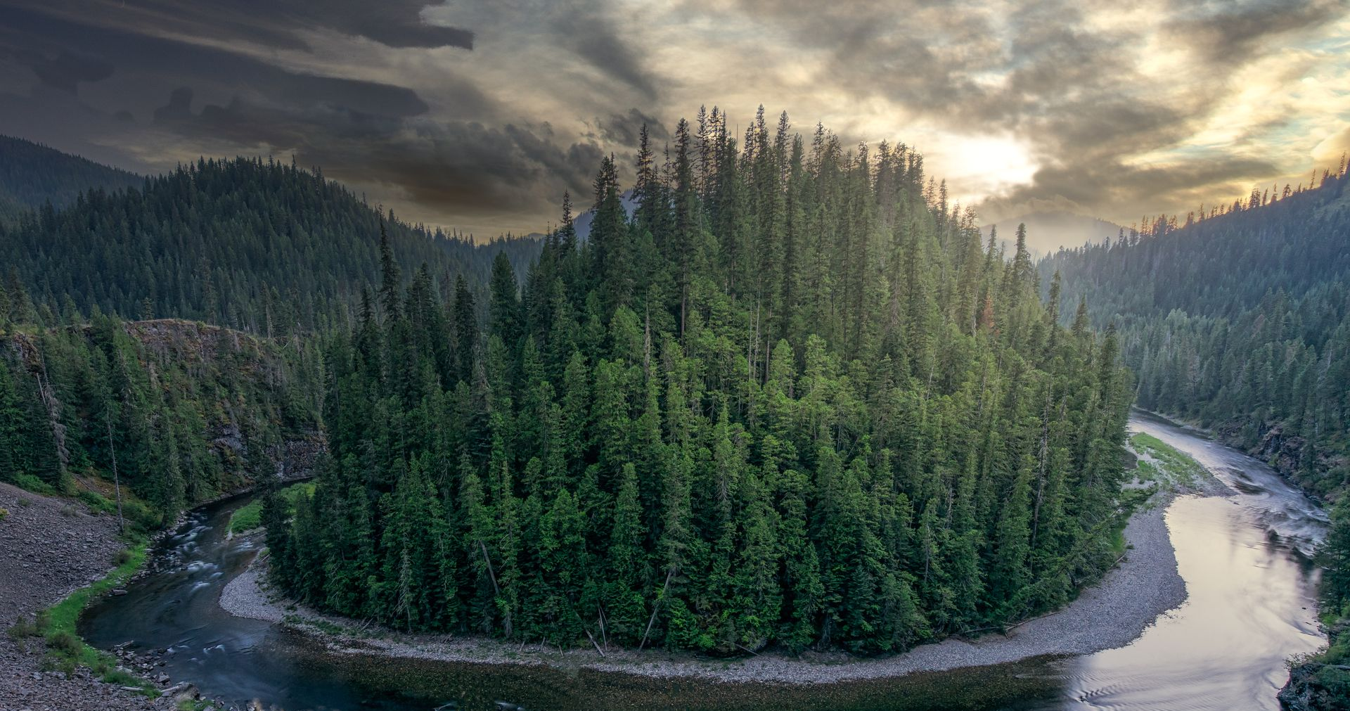 A winding river curves around a dense evergreen forest beneath a cloudy sunset sky.