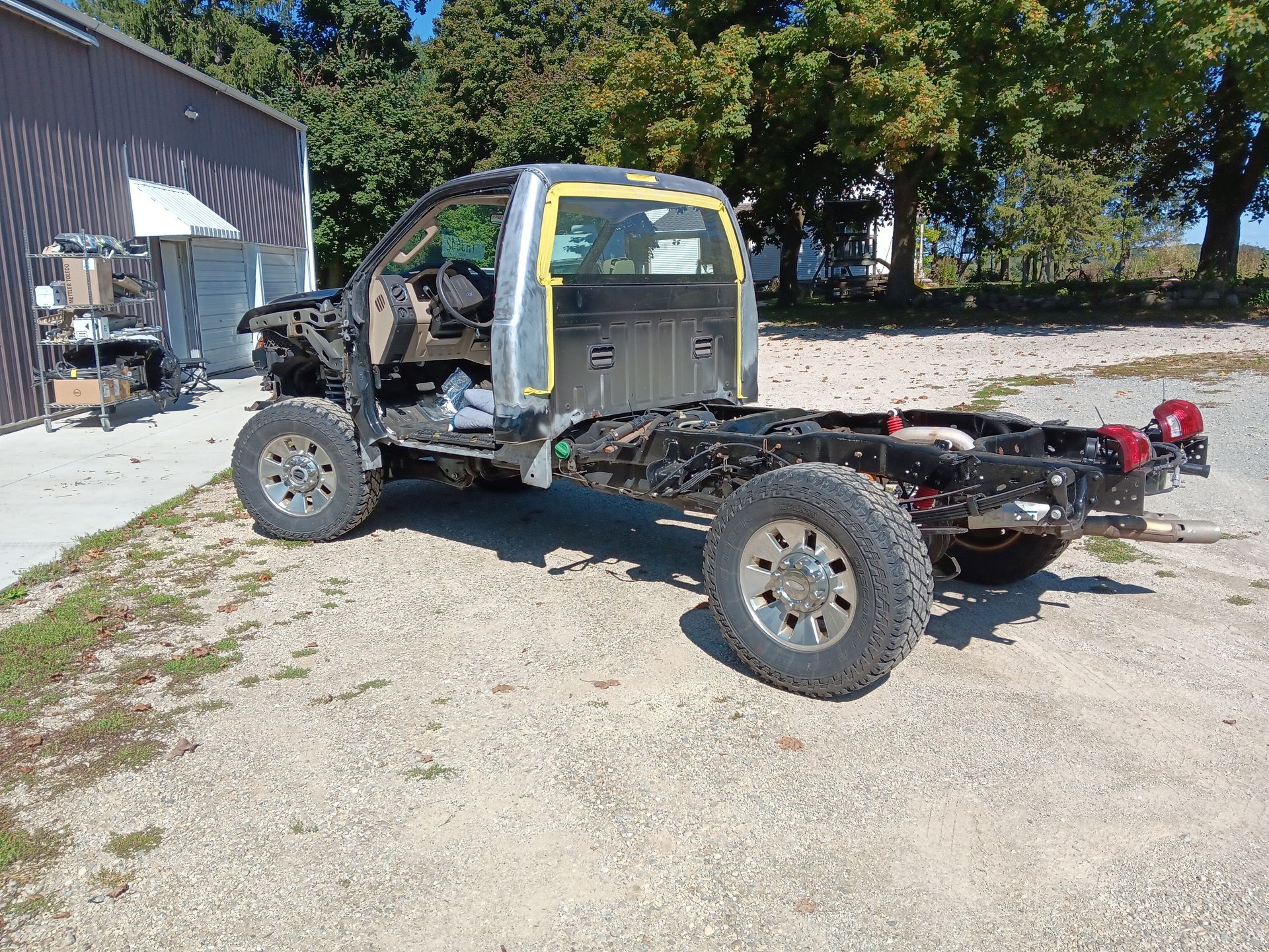 Truck chassis cab in a gravel yard beside a barn, with trees in the background.