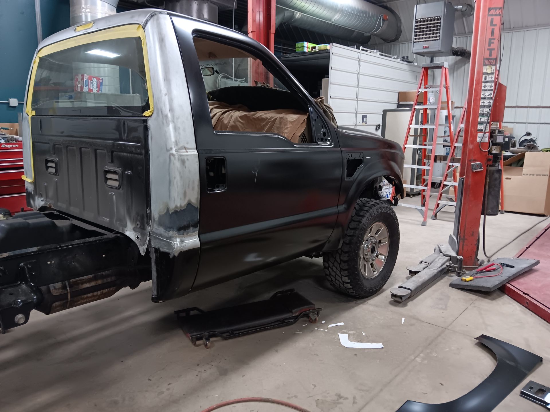 Black van in auto shop on lift, missing panels and front wheel, with tools and ladder nearby.