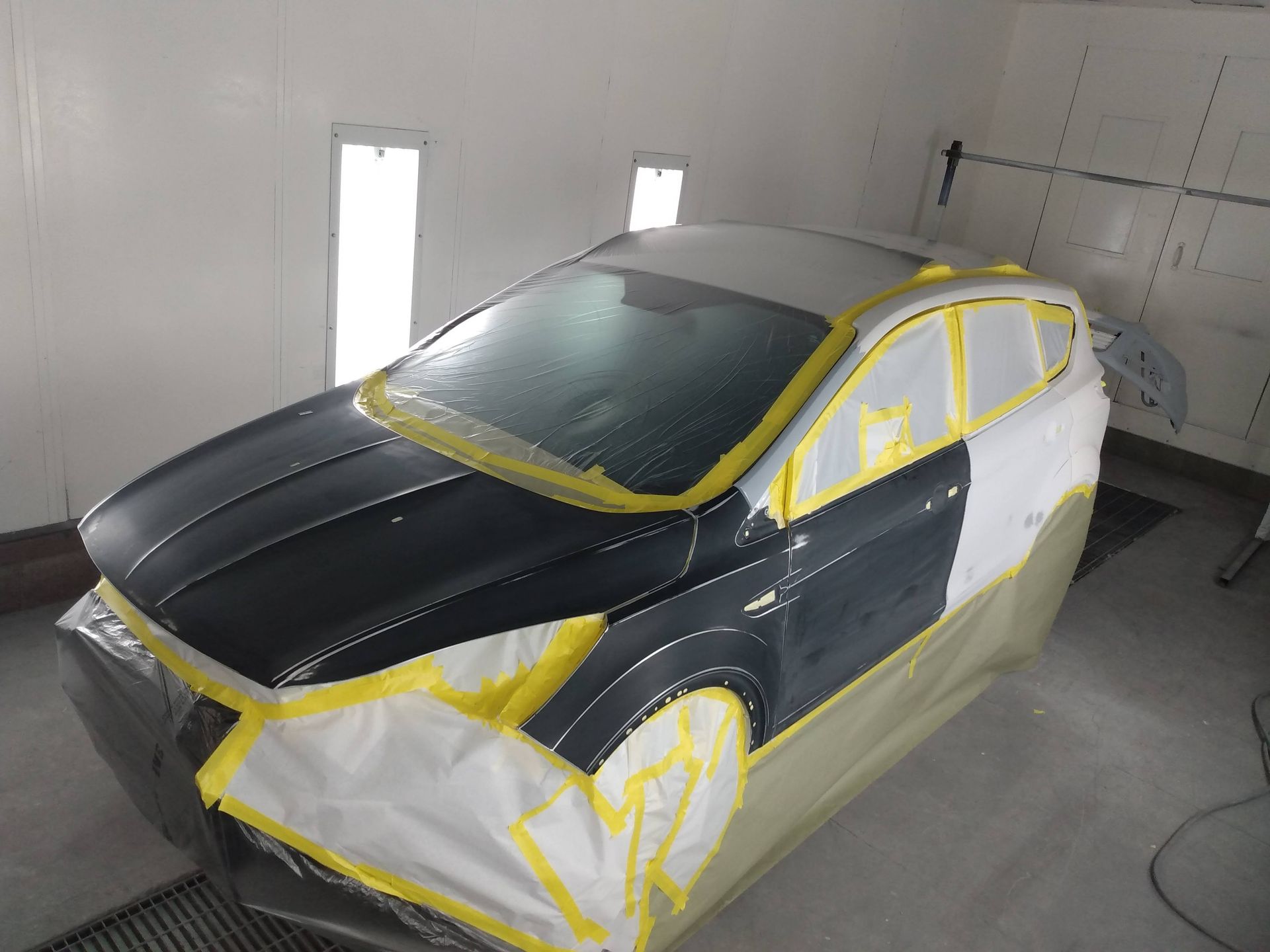 Car body in a paint booth, partially masked with yellow tape and plastic, black hood and roof.