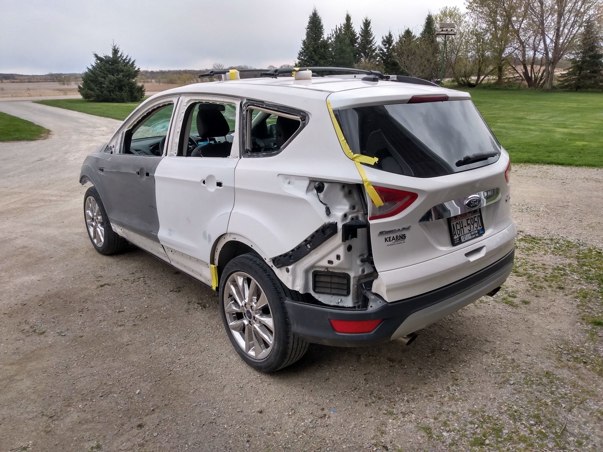 Damaged silver SUV with smashed rear and side windows parked on a gravel road by a grassy field.