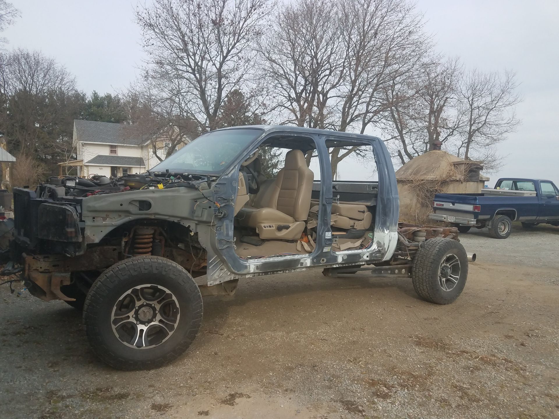 Damaged blue pickup truck with stripped body panels parked on a gravel lot under overcast sky