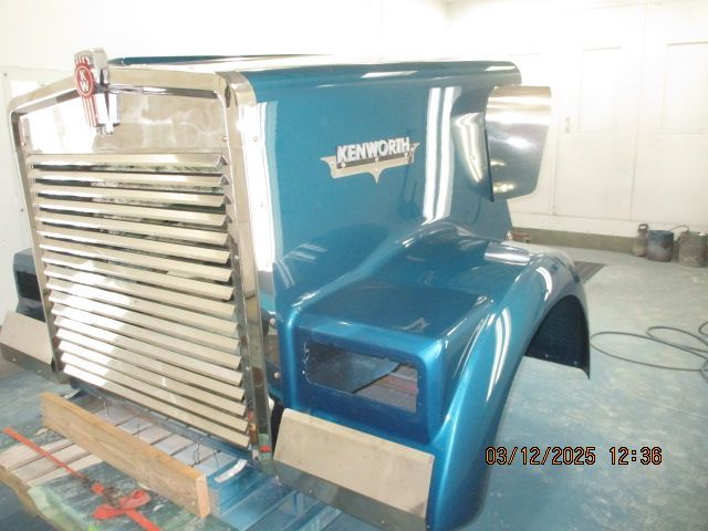 Blue Kenworth semi truck cab in a garage, front-left view with chrome grille and fender.