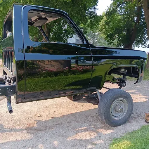 Glossy black lifted truck cab with no bed, parked on a gravel driveway under trees
