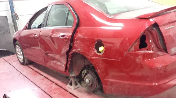 Red sedan with severe rear-side damage in a body shop, missing rear wheel and taillight.