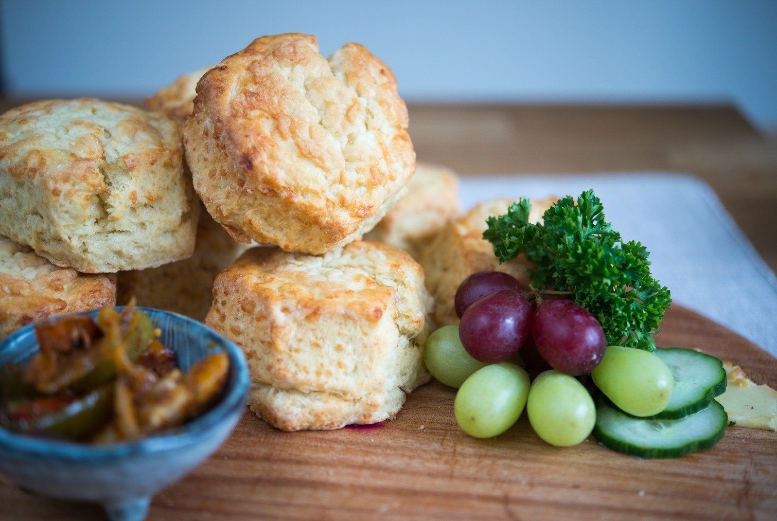 Biscuits piled on a wooden board with grapes, cucumber, parsley, and relish in a bowl.