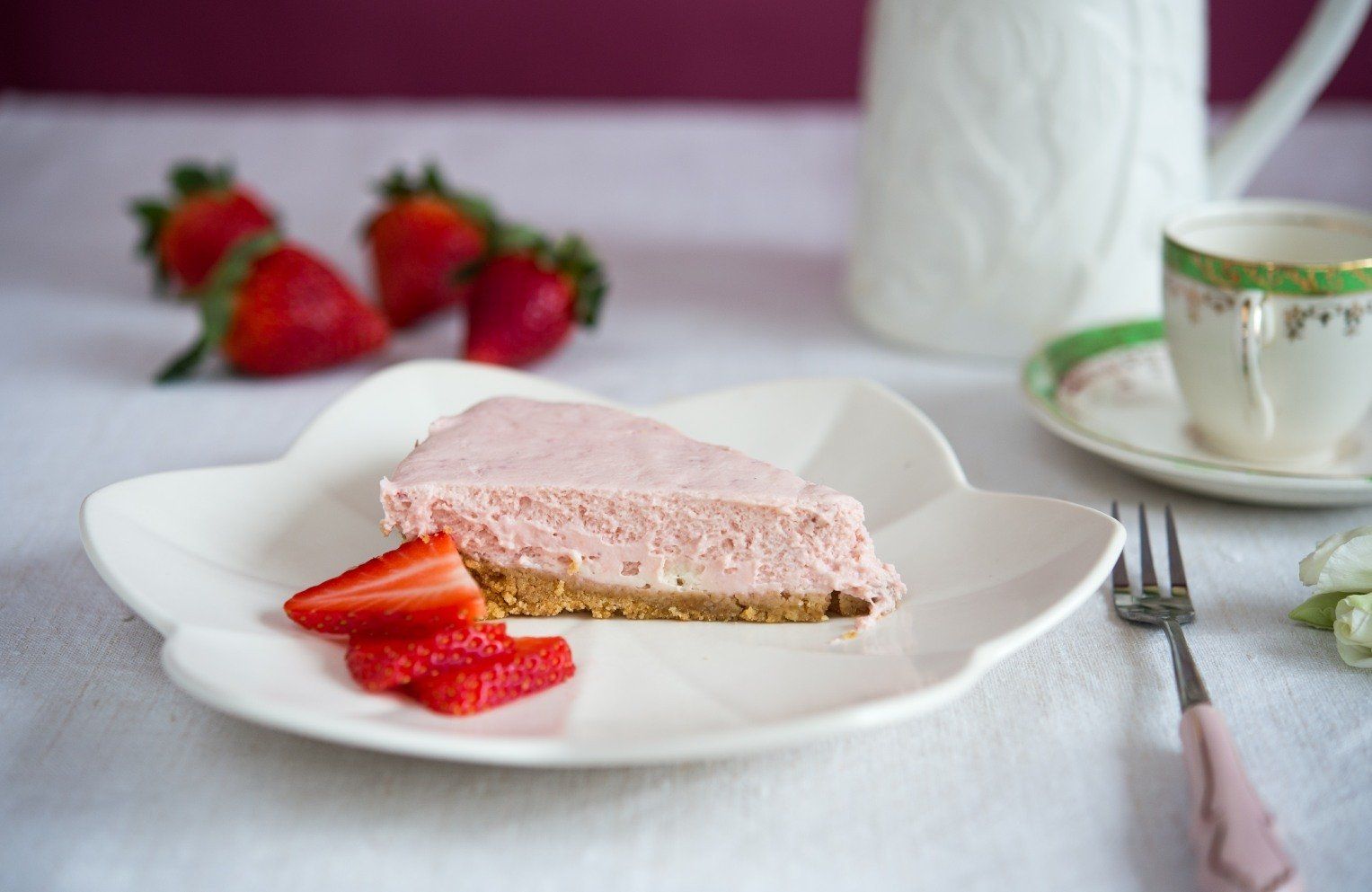 Slice of strawberry cheesecake on a flower-shaped plate with fresh strawberries, tea cup, and pitcher.
