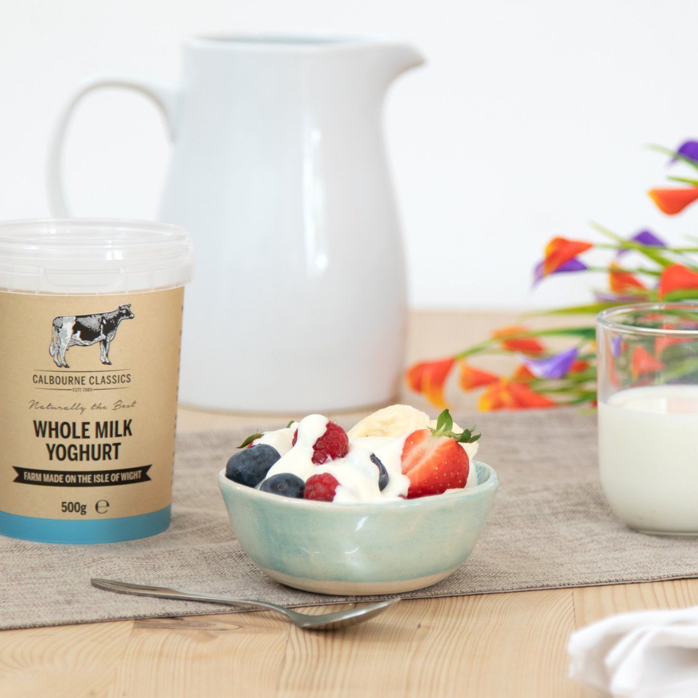 Yogurt bowl with berries, a jug, glass of milk, and yogurt container on a table.