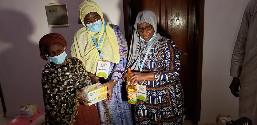 Three people stand indoors holding food items, including boxes and bottles.