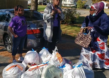Two women and a child stand by bags of donated food beside a parked car in a yard.
