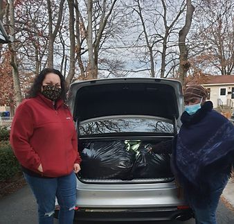 Two people stand beside an open car trunk filled with black bags in a wooded driveway.