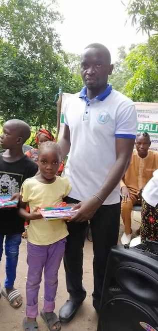 Man in white polo standing outdoors with children, one holding a small item, in a leafy area