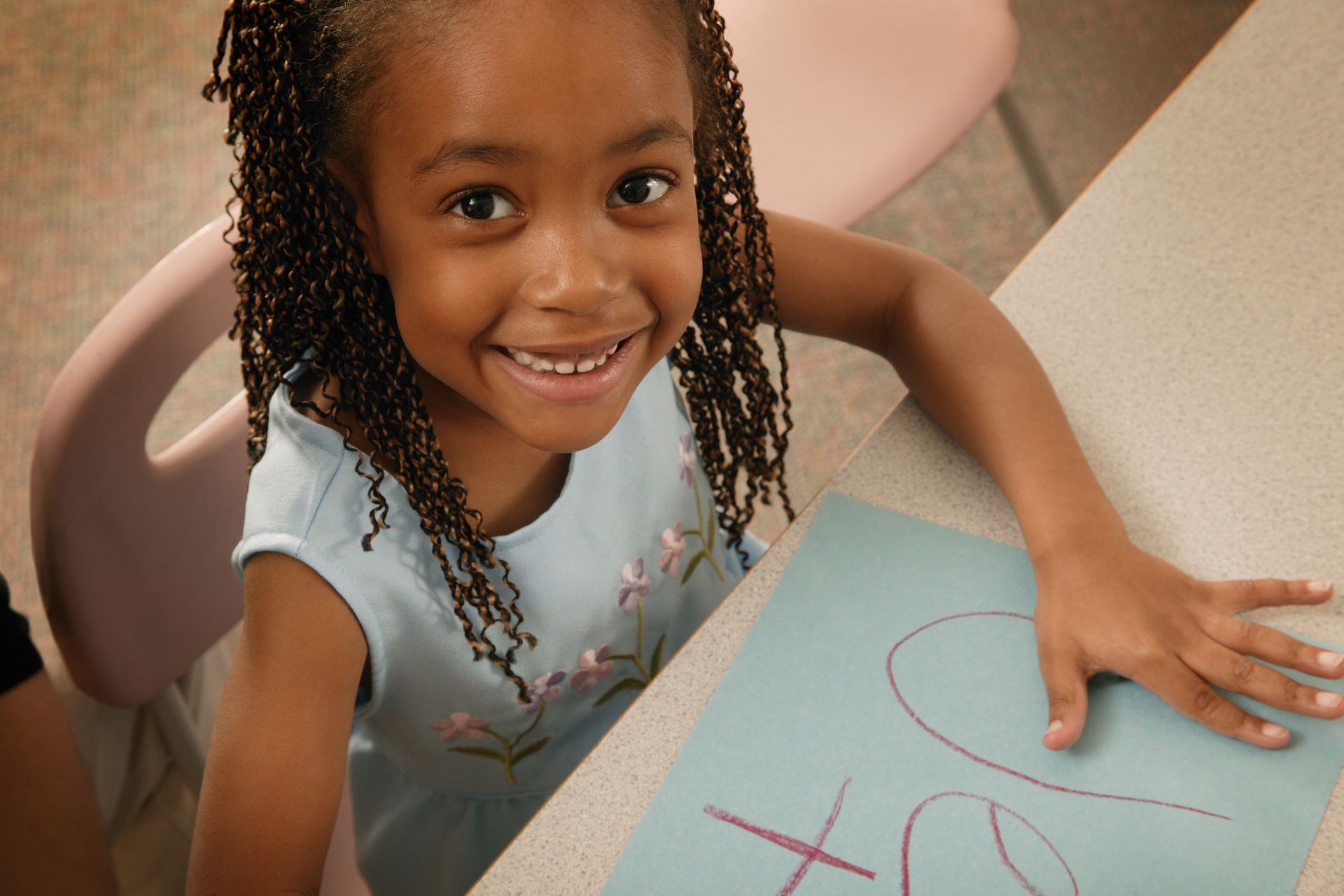 Smiling child at a table, writing on blue paper with a pink marker.