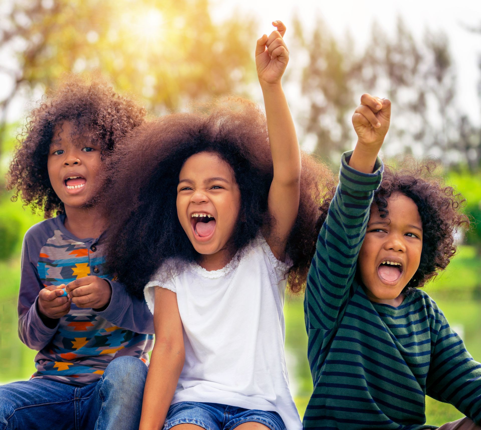 Three children cheering outdoors with raised arms in a sunny park
