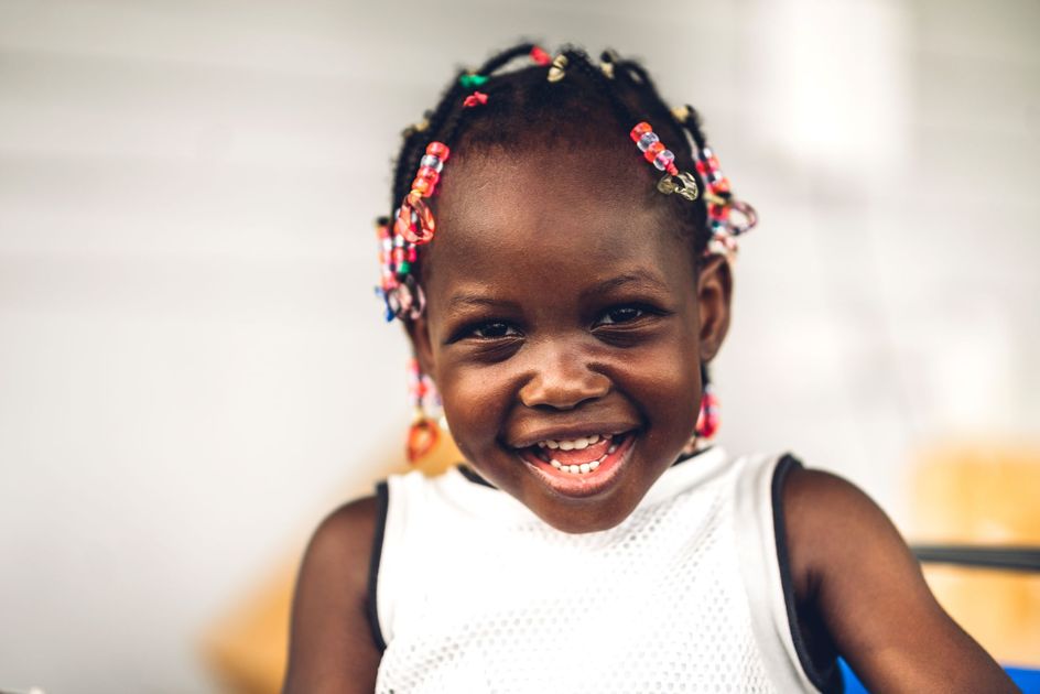 Smiling child in a white top with colorful beaded hair, against a softly blurred background