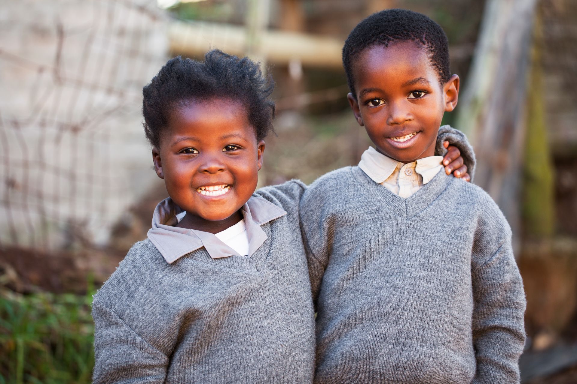 Two smiling children in gray sweaters standing outdoors with a blurred background
